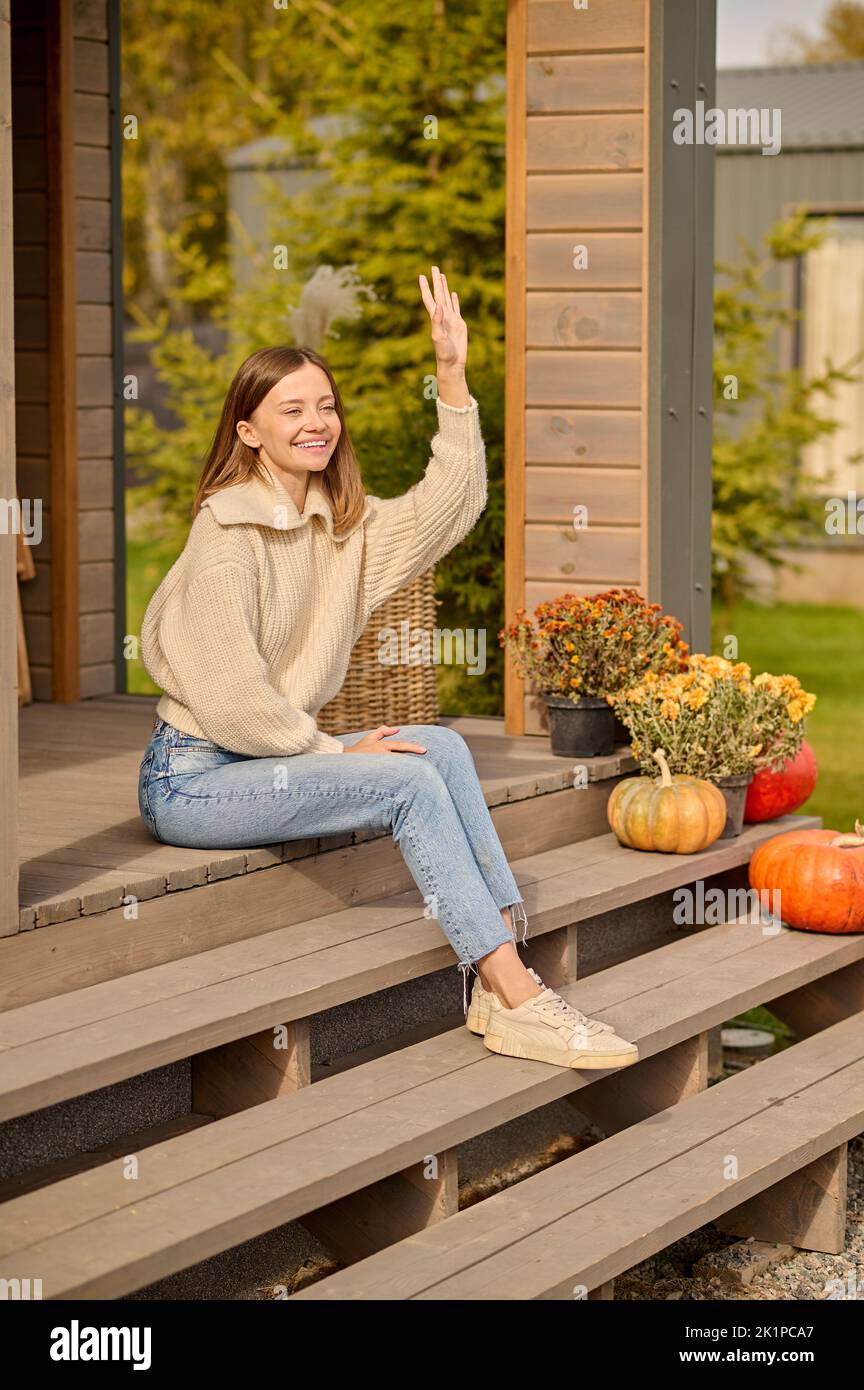 Joyous lady seated on the veranda stairs greeting someone Stock Photo