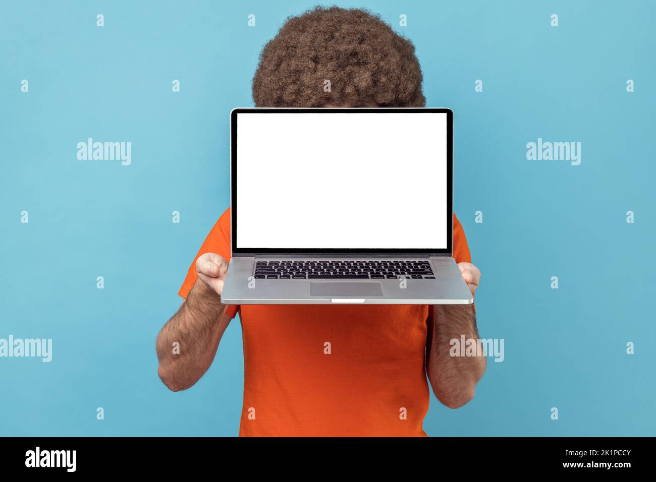 Unknown anonymous man with Afro hairstyle wearing orange T-shirt hiding his face with laptop with white empty display, copy space for promotional text. Indoor studio shot isolated on blue background. Stock Photo