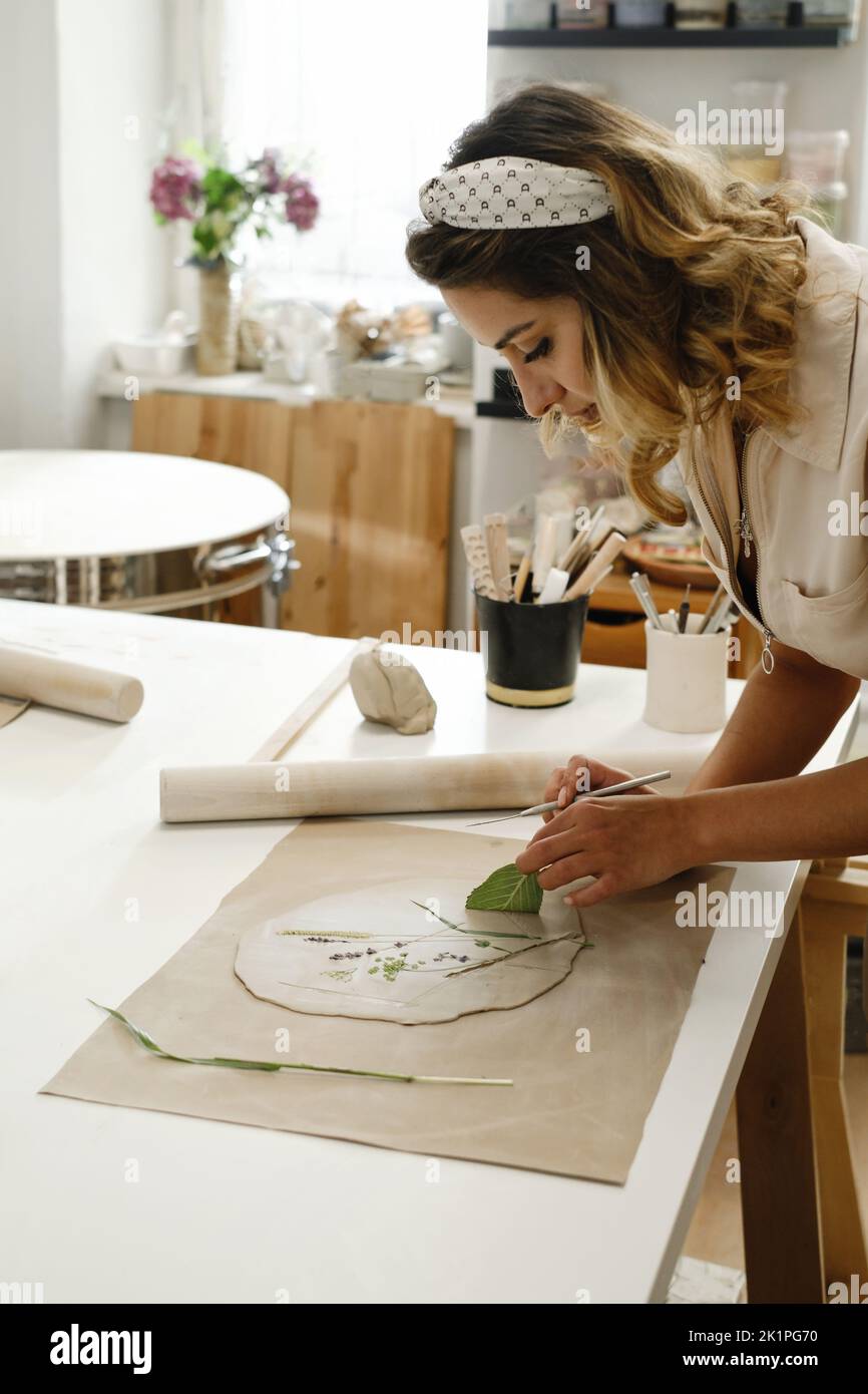 Woman decorating with flowers rolled clay, making ceramic plate in studio with floral pattern. Handmade creative work. Pottery workshop for adults.  Stock Photo