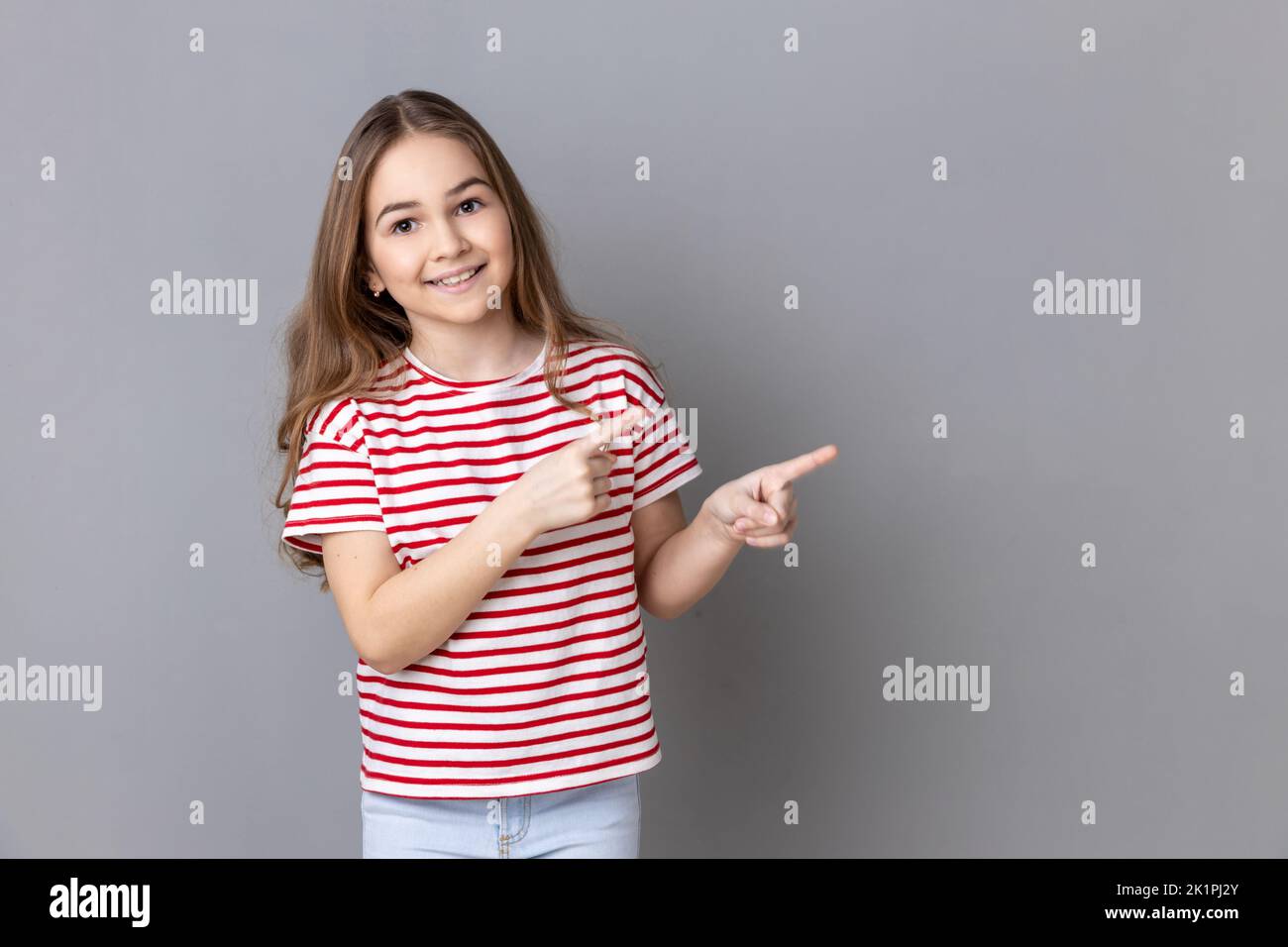 Attention to advertising. Little girl wearing striped T-shirt pointing aside, showing copy space for commercial text, blank wall with idea presentation. Indoor studio shot isolated on gray background. Stock Photo