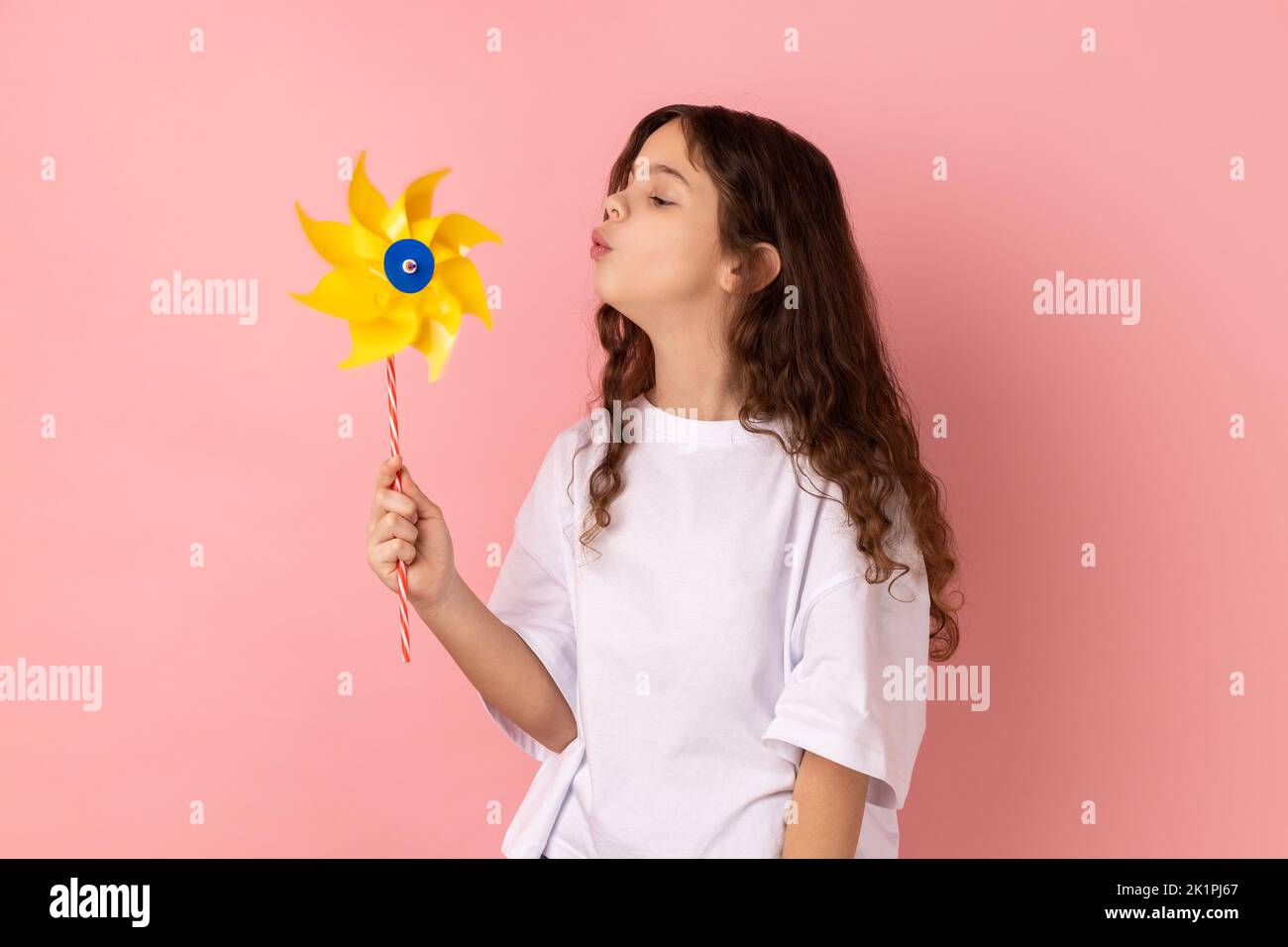 Portrait of charming cute playful little girl wearing white T-shirt blowing at paper windmill, playing with pinwheel toy on stick. Indoor studio shot isolated on pink background. Stock Photo