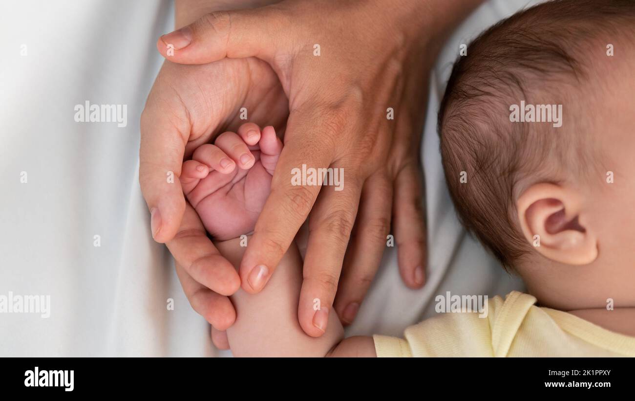 Young mother holding baby hand and showing finger and fingernail on white sheet bed. Closeup of baby sleeping on the mother's room. Family and home co Stock Photo