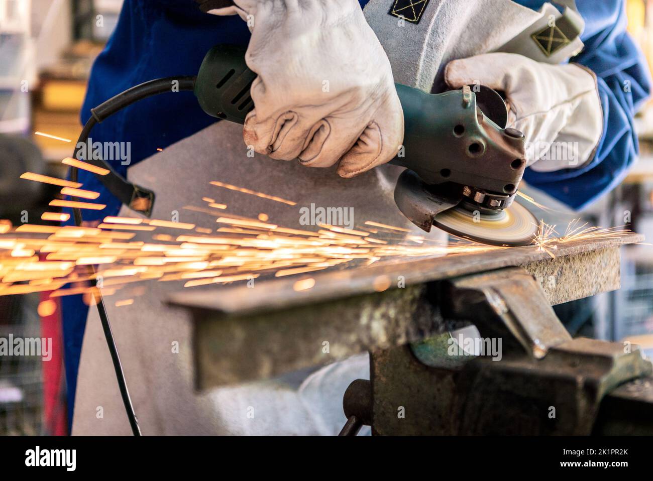Worker in special clothes and goggles grinding metal with a circular saw in his workshop, bright sparks of metalworking fly in different directions. Stock Photo