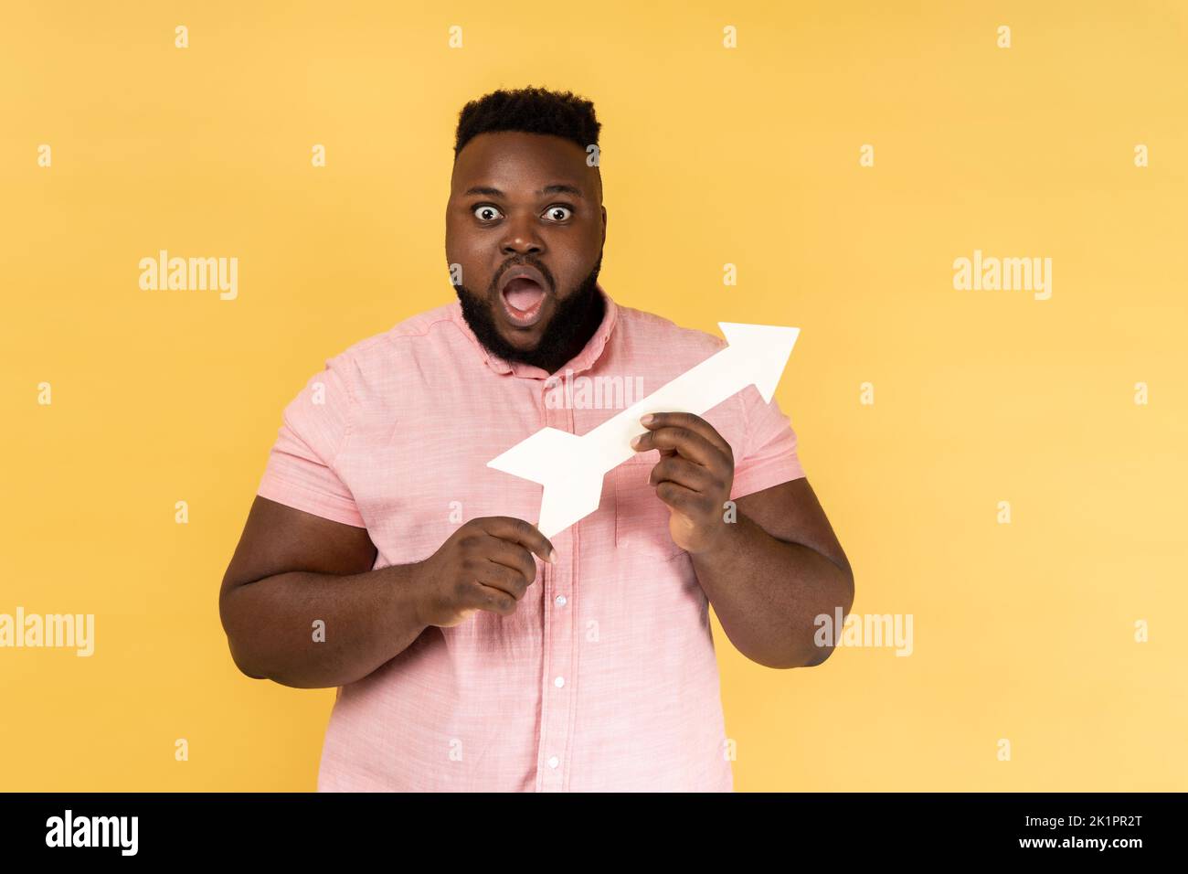 Portrait of astonished surprised man wearing pink shirt pointing direction with arrow aside, looking at camera with big eyes and open mouth. Indoor studio shot isolated on yellow background. Stock Photo