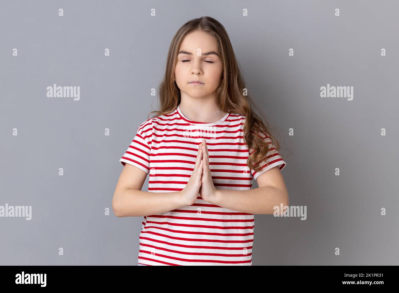 Portrait of little girl wearing striped T-shirt keeping hands namaste gesture, meditating, yoga exercise breath technique reduce stress. Indoor studio shot isolated on gray background. Stock Photo