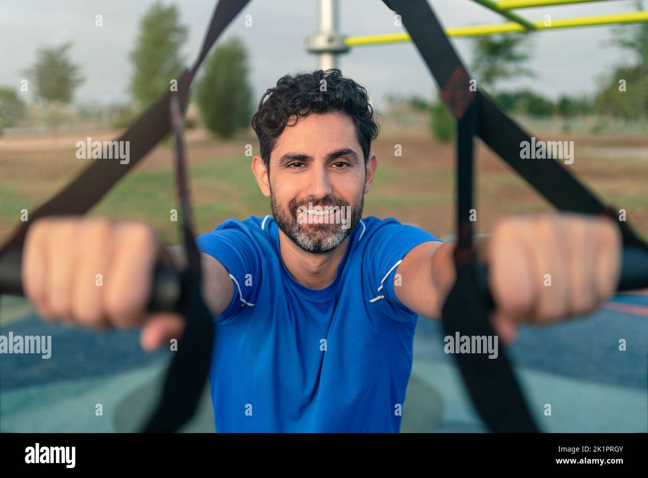 Portrait of confident sportsman working out with TRX system outside. Total body resistance exercises in city park. A handsome man is working out in st Stock Photo