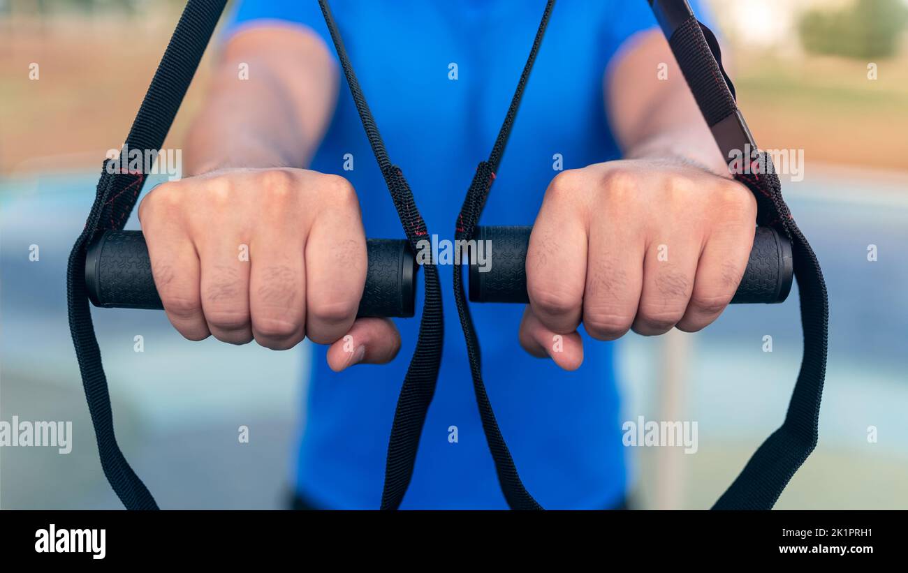 Close up of caucasian man hands exercising outside in sunny park. Great TRX workout. Young handsome guy in sportswear doing exercising outdoors. Healt Stock Photo