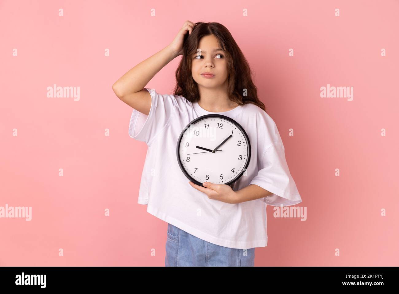 Portrait of confused puzzled little girl wearing white T-shirt holding wall clock and scratching her head, deadline, punctuality. Indoor studio shot isolated on pink background. Stock Photo