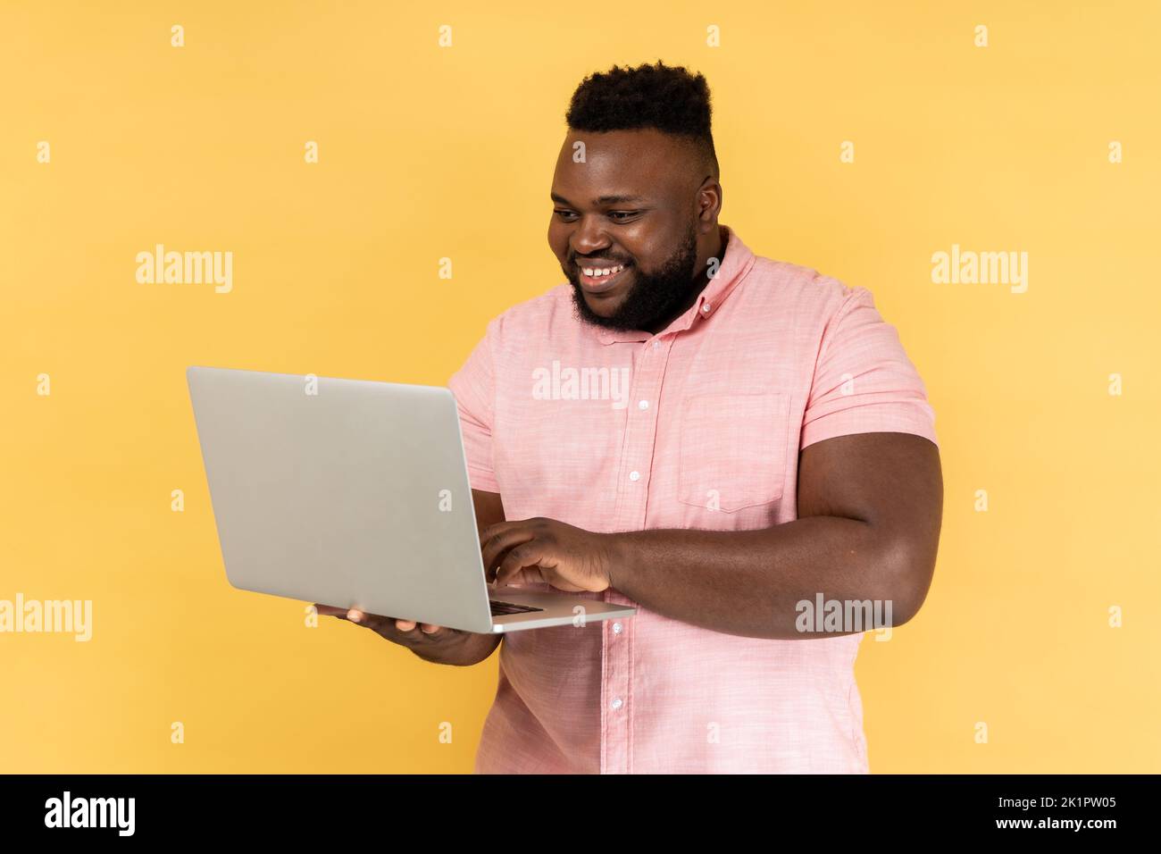 Portrait of positive freelancer man wearing pink shirt standing and holding laptop, satisfied with teleworking, likes his job, looking at display. Indoor studio shot isolated on yellow background. Stock Photo