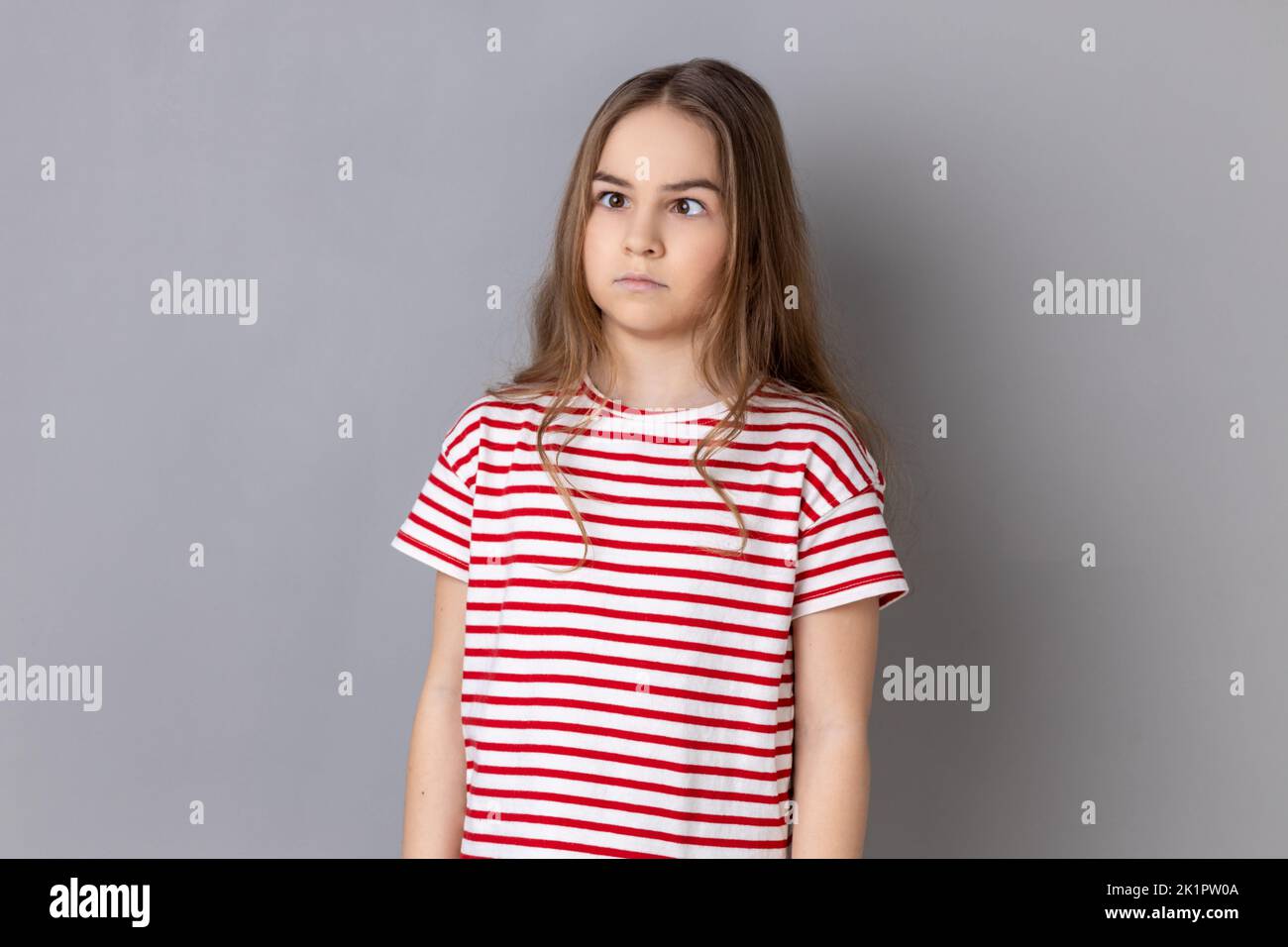 Portrait of funny silly little girl wearing striped T-shirt looking up cross eyed with stupid dumb face, girl has awkward confused comical expression. Indoor studio shot isolated on gray background. Stock Photo