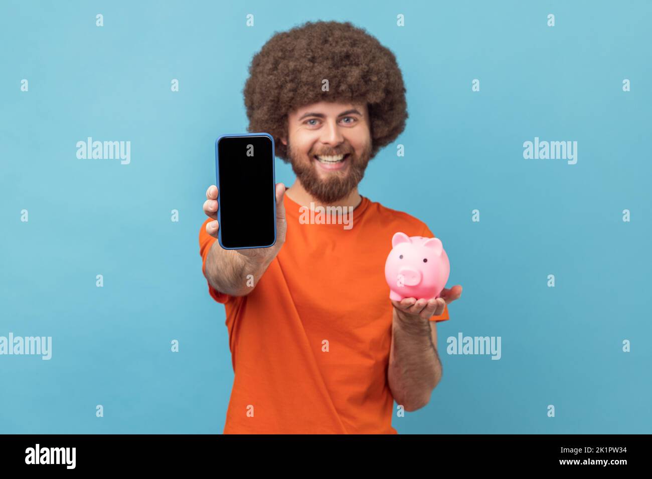 Portrait of happy satisfied man with Afro hairstyle in orange T-shirt holding in hands piggy bank and smart phone with empty screen for advertisement. Indoor studio shot isolated on blue background. Stock Photo