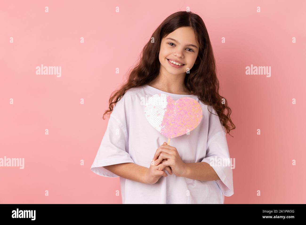 Portrait of happy satisfied little girl wearing white T-shirt holding paper pink heart on stick and smiling to camera, sharing love. Indoor studio shot isolated on pink background. Stock Photo