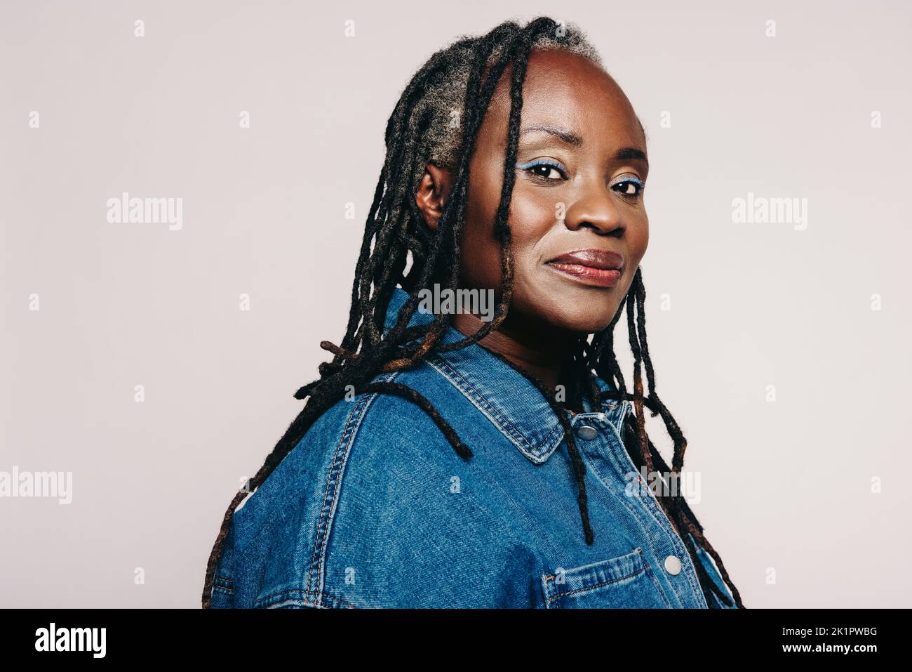 Stylish woman with dreadlocks looking at the camera while standing against a grey background. Portrait of a mature woman wearing a denim jacket and ma Stock Photo