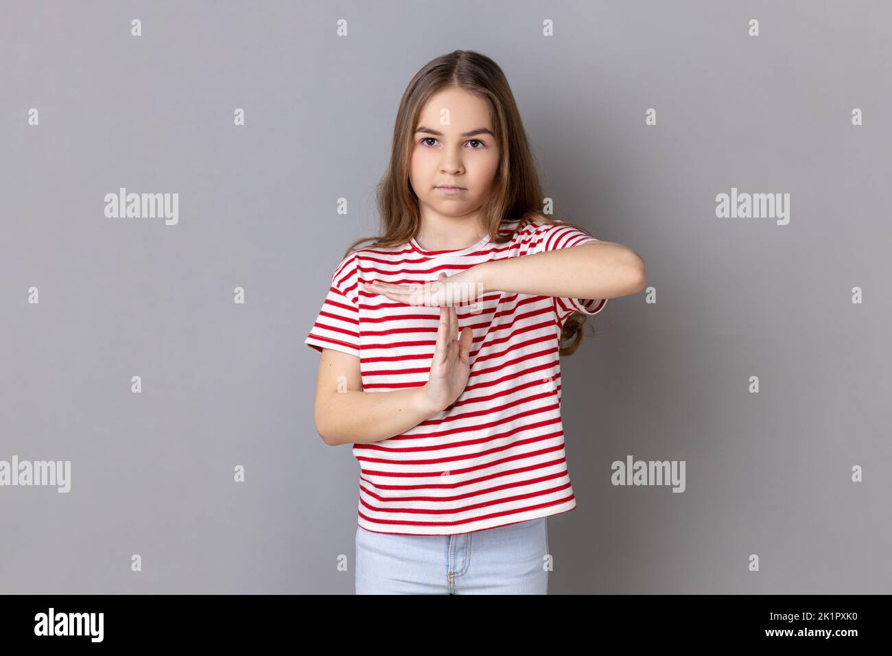 Portrait of frustrated little girl wearing striped T-shirt showing time out gesture, looking at camera, deadline with test in school. Indoor studio shot isolated on gray background. Stock Photo
