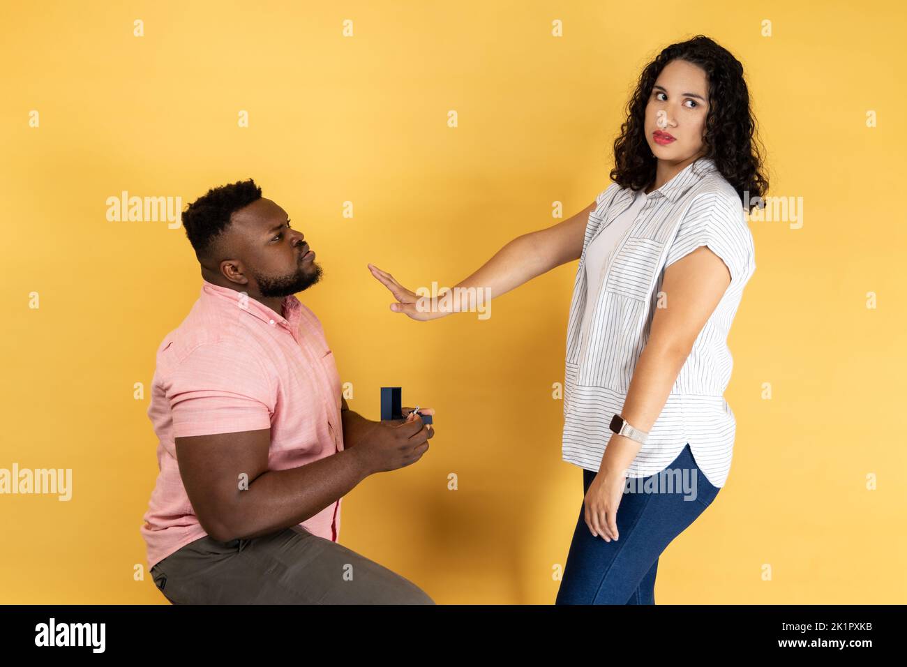 Portrait of young couple in casual clothing, man making a proposal to his girlfriend, woman refusing, showing stop palm gesture. Indoor studio shot isolated on yellow background. Stock Photo