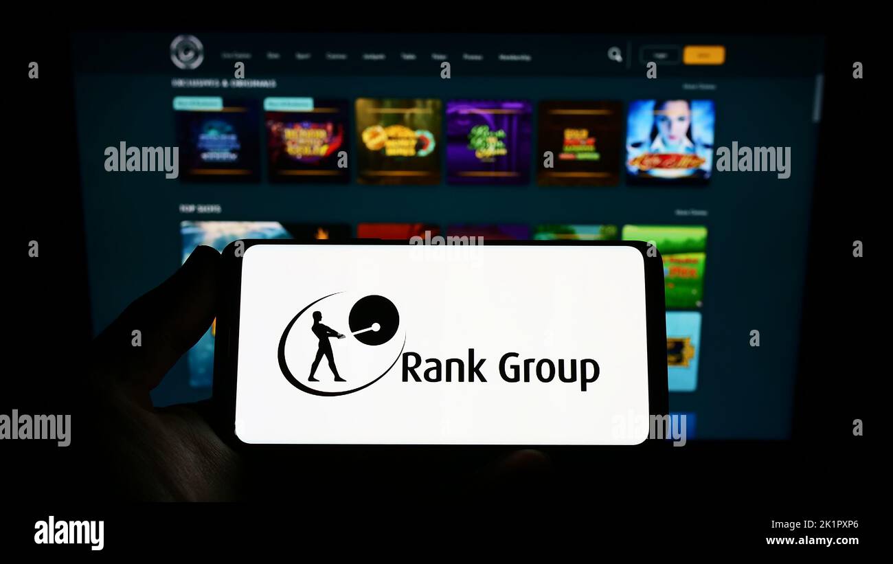 Person holding smartphone with logo of British gambling company The Rank Group plc on screen in front of website. Focus on phone display. Stock Photo