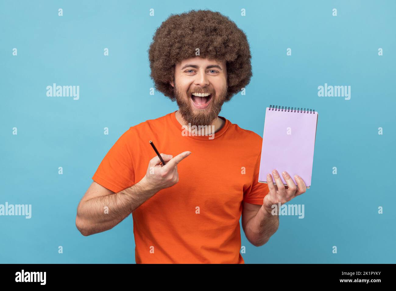 Write reminder to-do list into notebook. Man with Afro hairstyle showing empty paper page, template mock up for creative idea, advertising text. Indoor studio shot isolated on blue background. Stock Photo