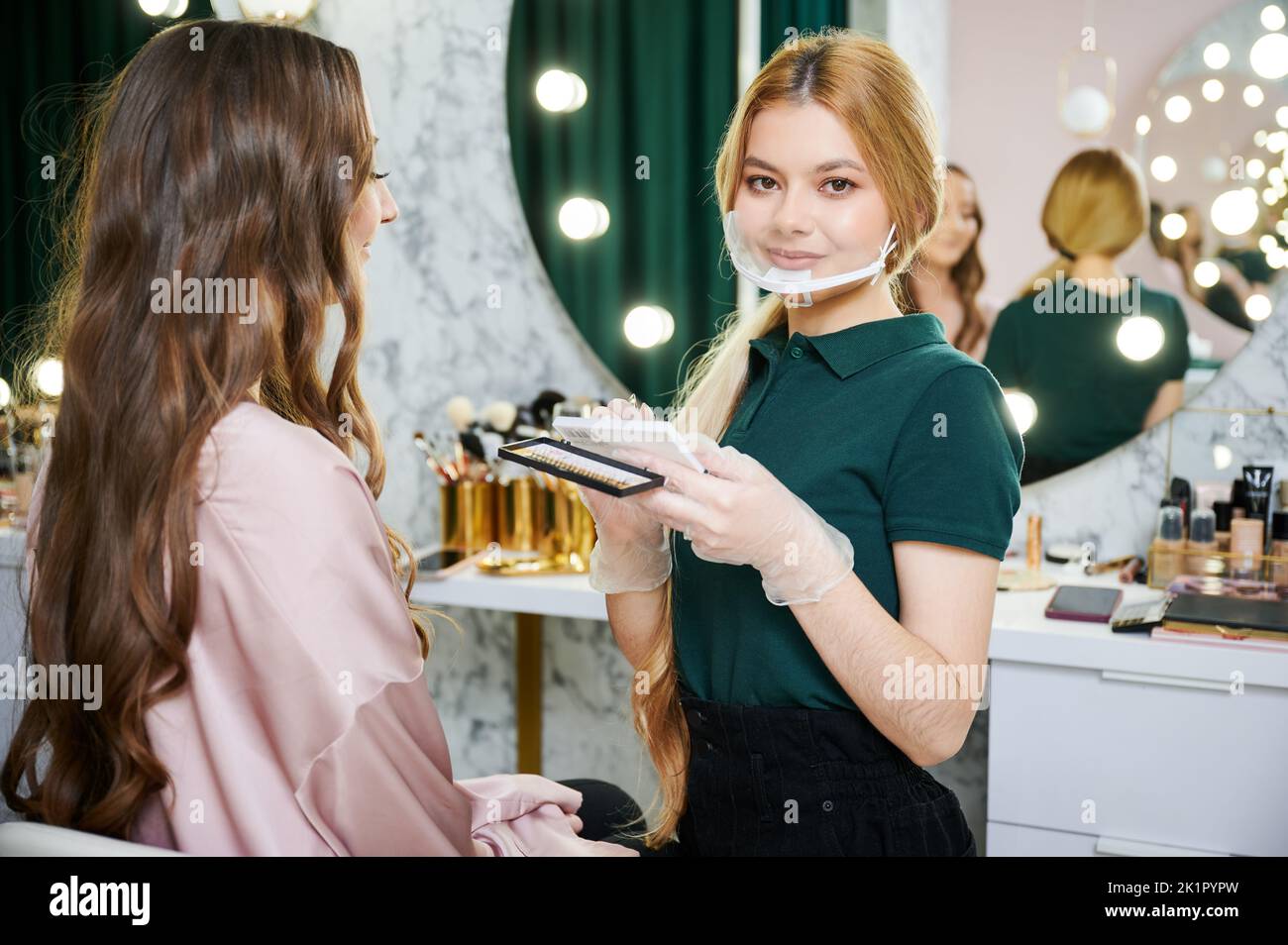 Portrait of beauty specialist holding eyelashes set while standing near young woman in visage studio. Female makeup artist in face mask looking at camera while client sitting on chair in beauty salon. Stock Photo