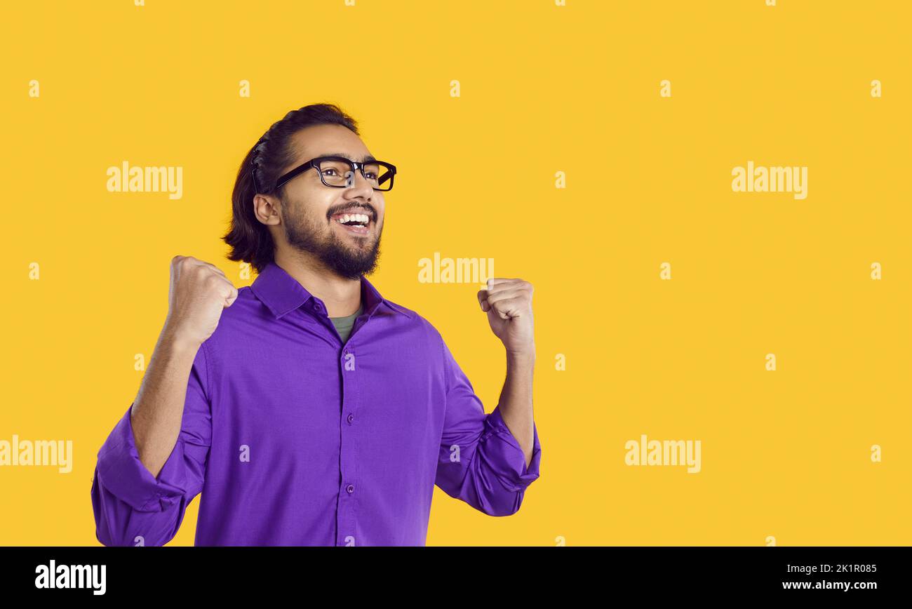 Smiling young South Asian man raises hands as sign successful, standing in yellow studio Stock Photo