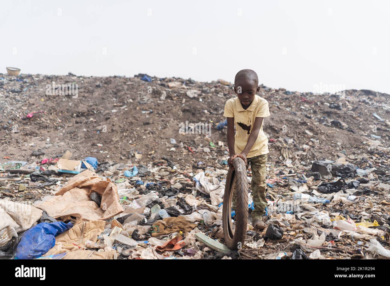 Underweight African boy playing with his bicycle wheel amid plastic and toxic substances at a landfill; risk of stunting and disease transmisison in d Stock Photo