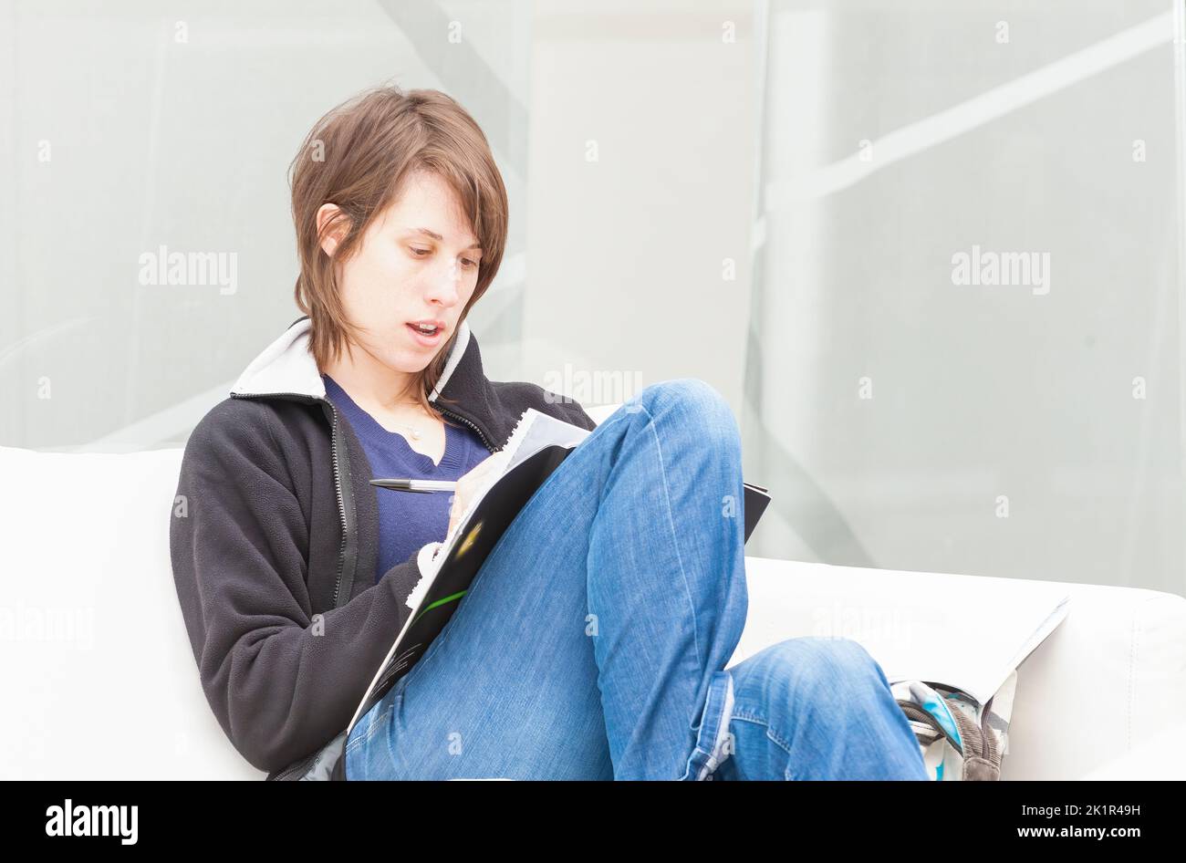 A casually dressed young woman, sitting on a white sofa and reading from and writing in, a book Stock Photo