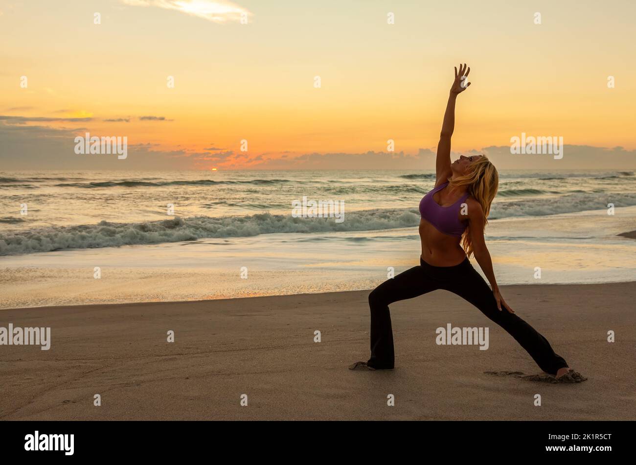 Young woman girl female in a warrior position practicing yoga on a beach at sunrise or sunset Stock Photo
