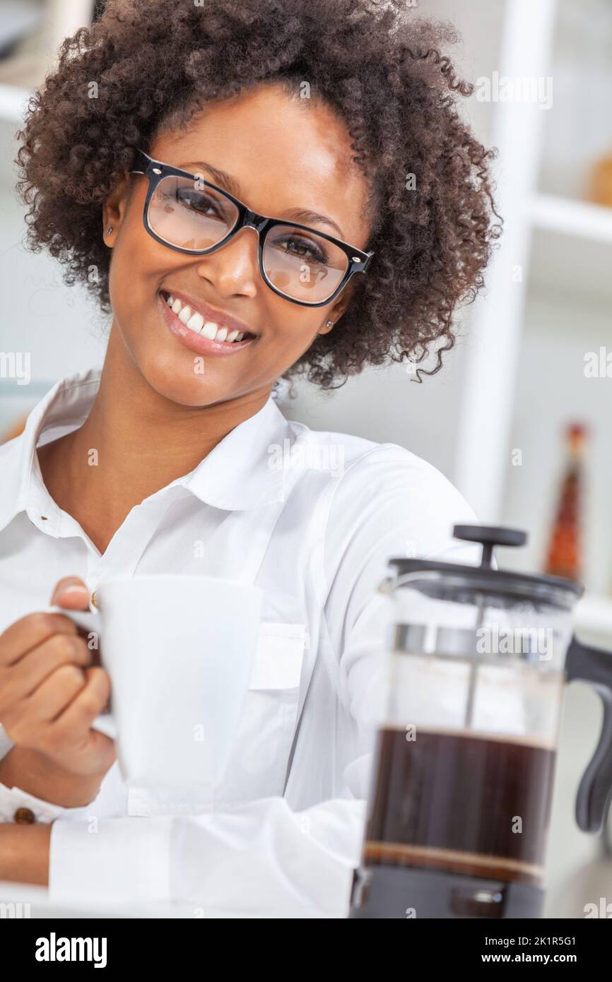 Beautiful happy biracial African American girl or young woman with perfect teeth wearing glasses in her kitchen, drinking a mug of coffee Stock Photo