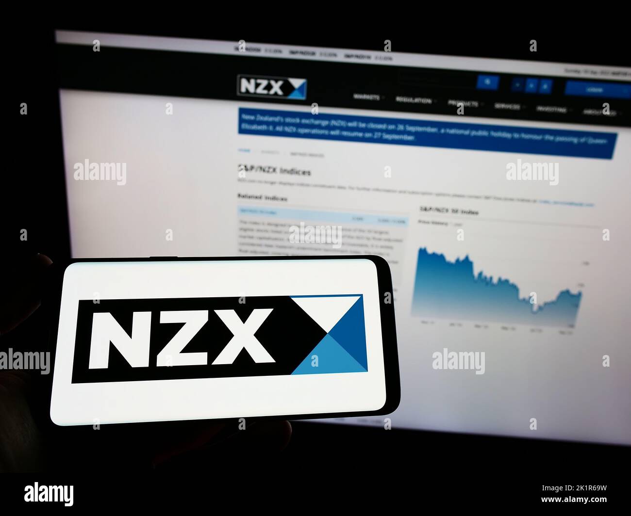Person holding mobile phone with logo of financial company New Zealand's Exchange (NZX) on screen in front of web page. Focus on phone display. Stock Photo