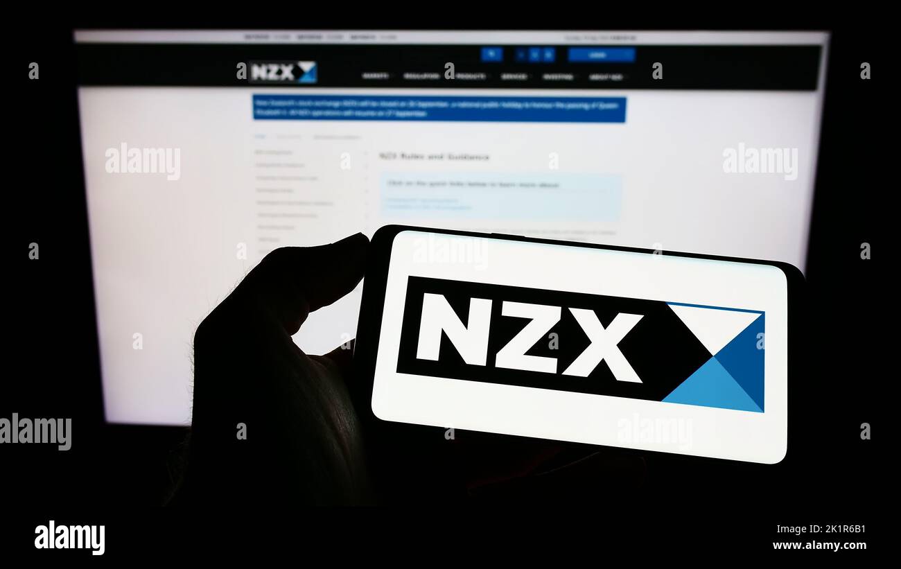 Person holding cellphone with logo of financial company New Zealand's Exchange (NZX) on screen in front of webpage. Focus on phone display. Stock Photo