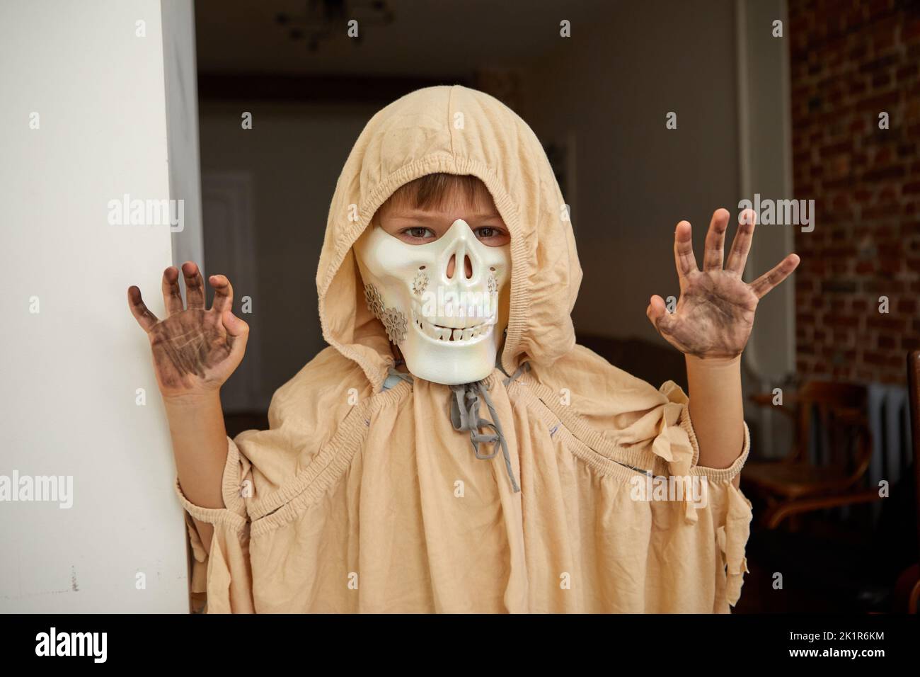 Child in spooky Halloween cape and skull jaw mask showing frightening gesture while looking at camera at home and having fun during holiday celebratio Stock Photo