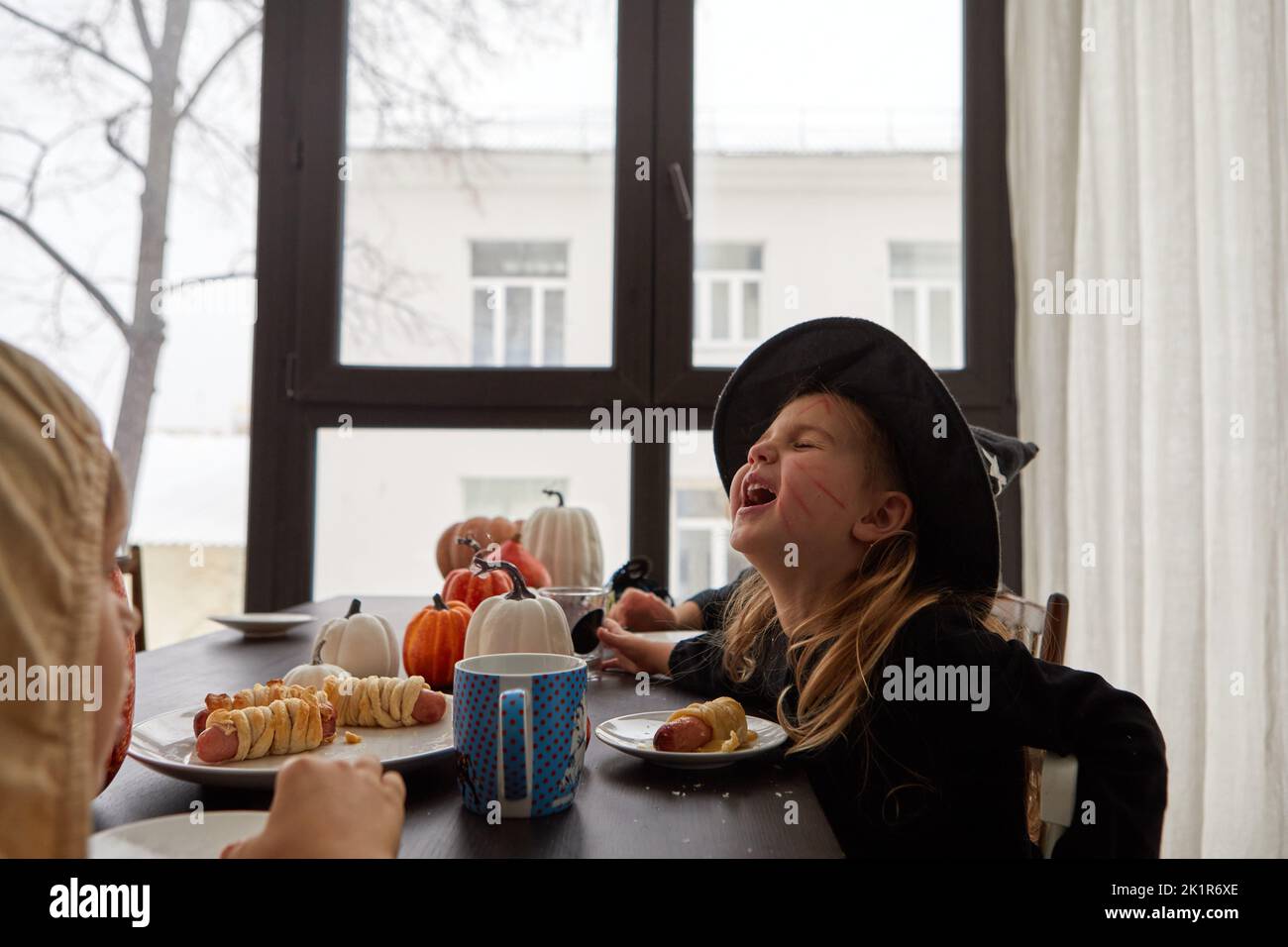 Cheerful child in costume of witch sitting at table and eating sausage roll while celebrating Halloween holiday at home and having fun Stock Photo