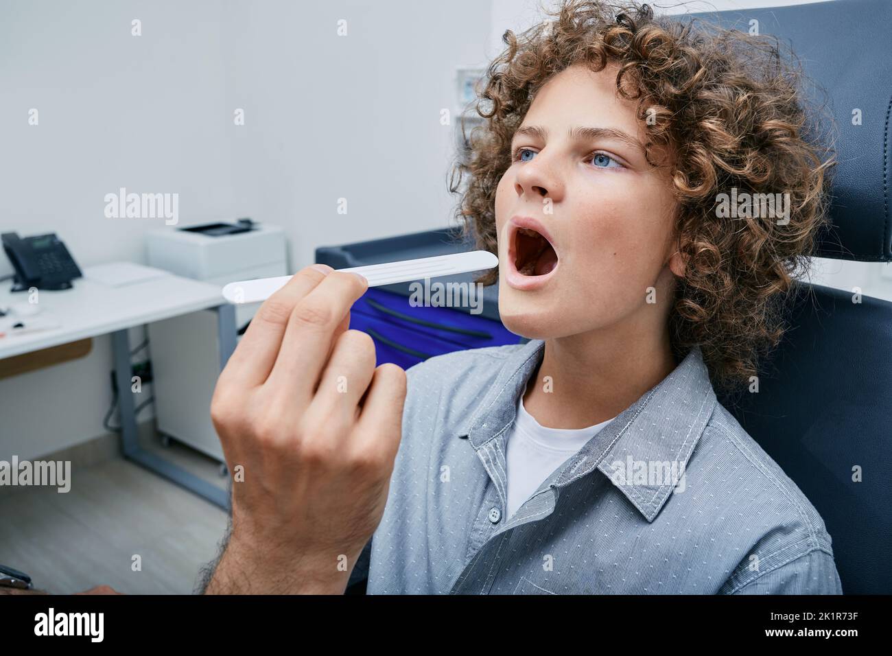 Curlyhaired boy opens his mouth during throat examination using tongue depressor by doctor at