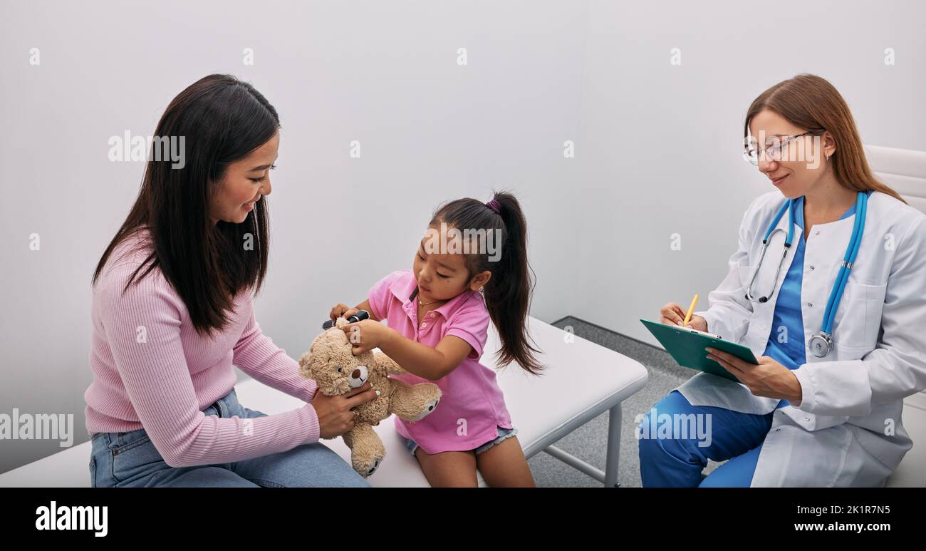 Asian girl plays with her toy bear in medical game, using otoscope with support of her mom and audiologist. Hearing clinic for children Stock Photo