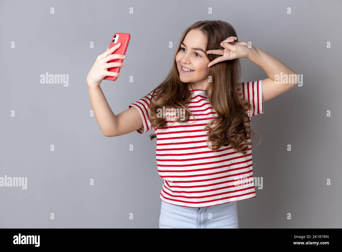Portrait of little girl wearing striped T-shirt makes peace sign or V gesture at camera of cell phone, poses for making selfie or video call. Indoor studio shot isolated on gray background. Stock Photo