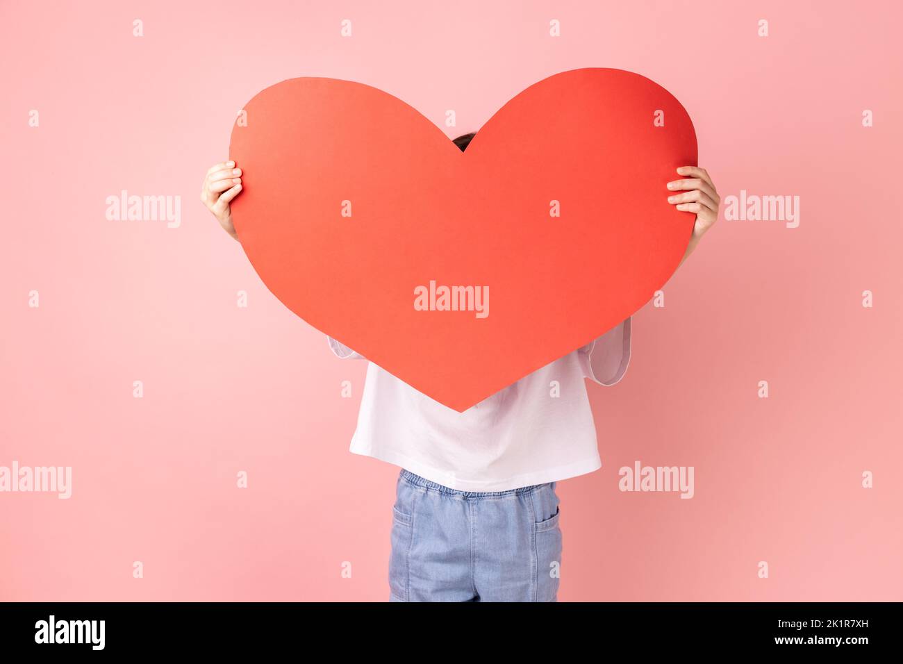 Portrait of anonymous little girl wearing white T-shirt hiding his face behind big red paper heart, making anonymous surprise, sending greeting card. Indoor studio shot isolated on pink background. Stock Photo