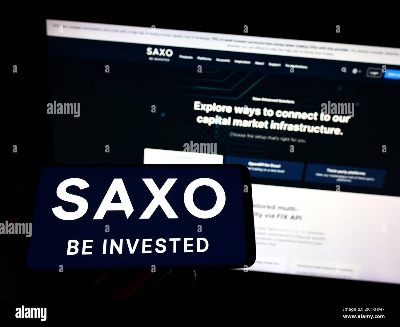 Person holding mobile phone with logo of Danish investment banking company Saxo Bank AS on screen in front of web page. Focus on phone display. Stock Photo