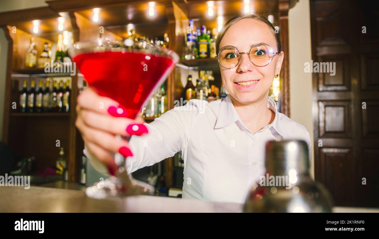 Female bartender in eyeglasses with cocktail standing behind bar, looking at camera, offering alcoholic beverage, smiling Stock Photo