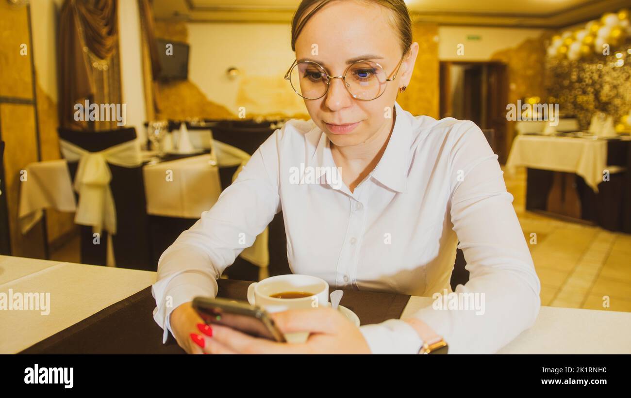 Young woman in glasses drinking tea, browsing mobile phone, sitting alone in restaurant in evening Stock Photo