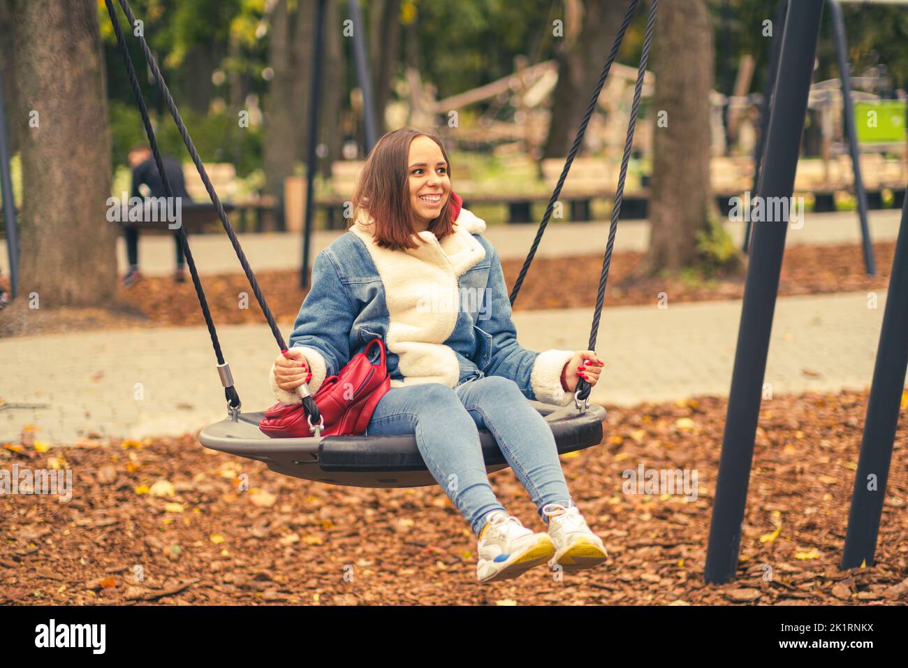 A beautiful and cheerful woman in warm clothes swings on a swing in the park. Girl swinging in the autumn city park. Stock Photo