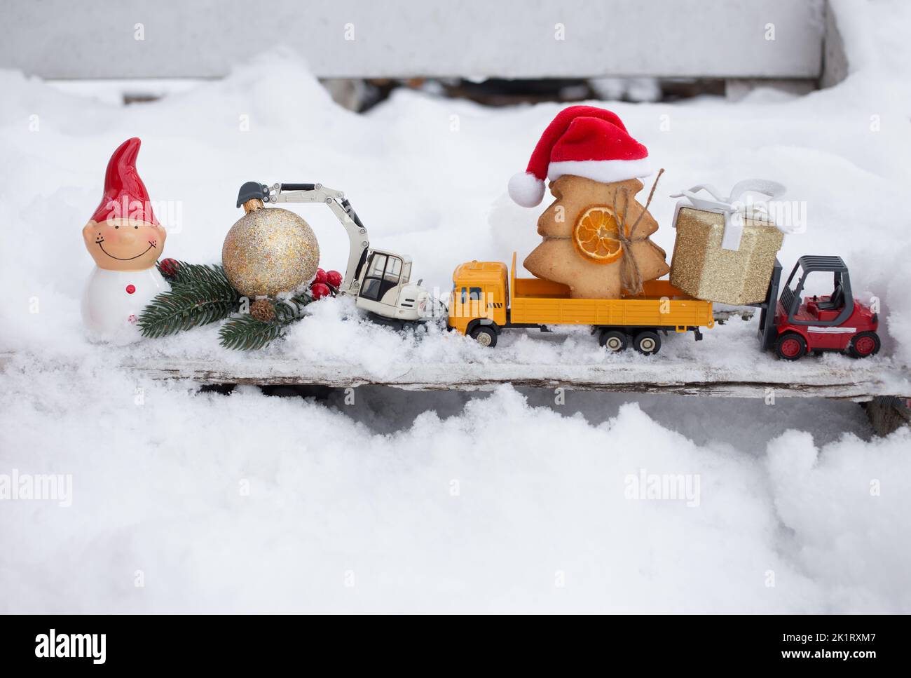 model of toy metal excavator, truck loaded with gingerbread tree, decorative Santa's helper snowman, warehouse loader standing on snow. concept of Chr Stock Photo