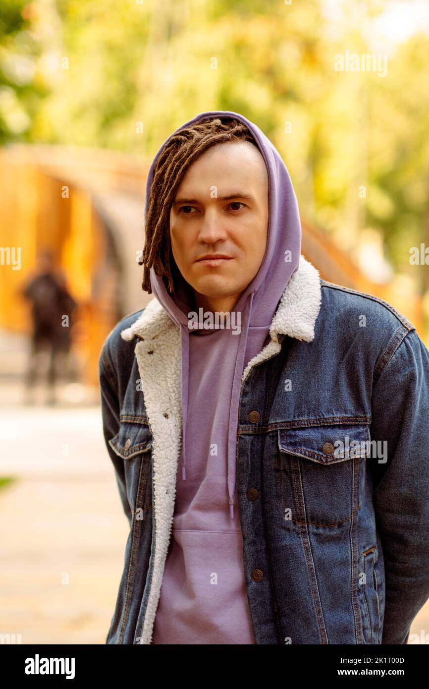 Portrait of handsome man with brown dreadlocks in hoodie, denim jacket posing on street in city park Stock Photo