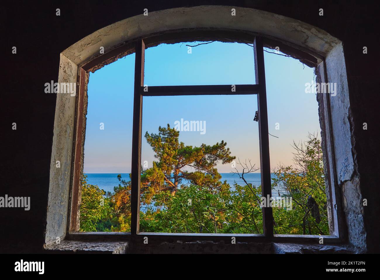 View through an old cracked window to the sea and trees illuminated by the setting sun. Stock Photo