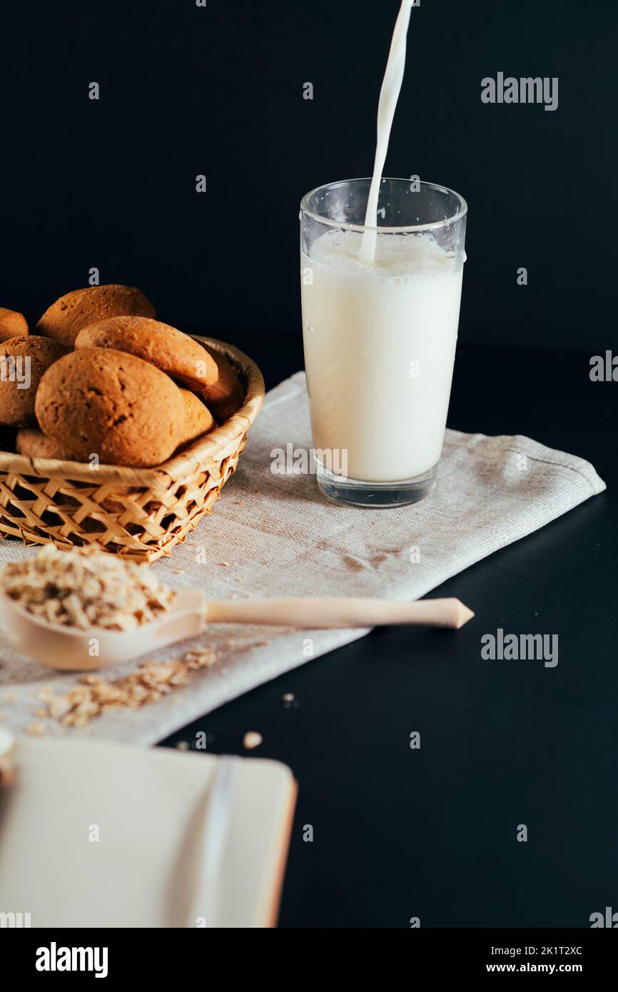 Basket of fresh oatmeal cookies with raw oat flakes in large wooden spoon on napkin on black background. Unrecognizable person pouring milk into glass Stock Photo