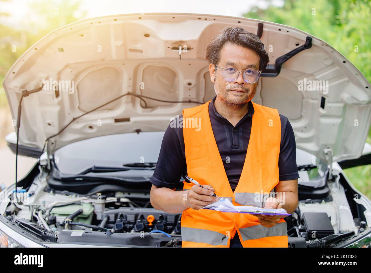 male in safety uniform standing with check list at car open front hood. claim adjuster engineer mechanic man portrait happy. Stock Photo