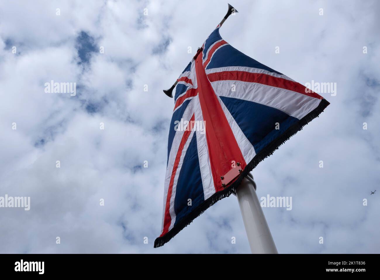 British national flag like a banner on a pole fluttering in the wind against a cloudy sky. Plane in background Stock Photo