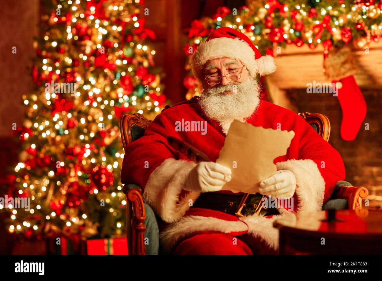 Portrait of traditional Santa Claus reading letter by fireplace on Christmas eve and smiling, copy space Stock Photo
