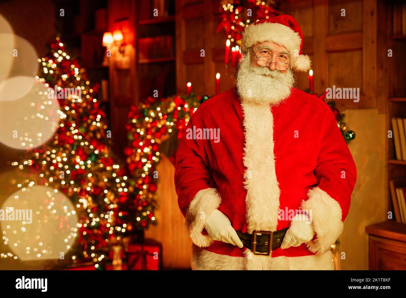 Waist up portrait of traditional Santa Claus looking at camera while standing in room with Christmas tree and twinkling lights, copy space Stock Photo