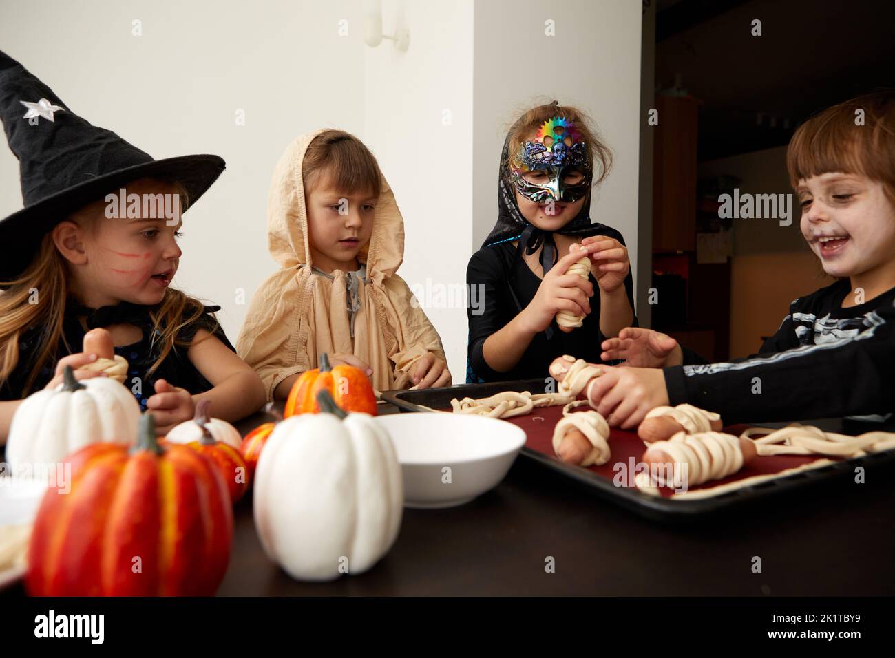 Company of content friendly kids in Halloween costumes gathering at table and prepare cooking sausage rolls during holiday celebration at home Stock Photo