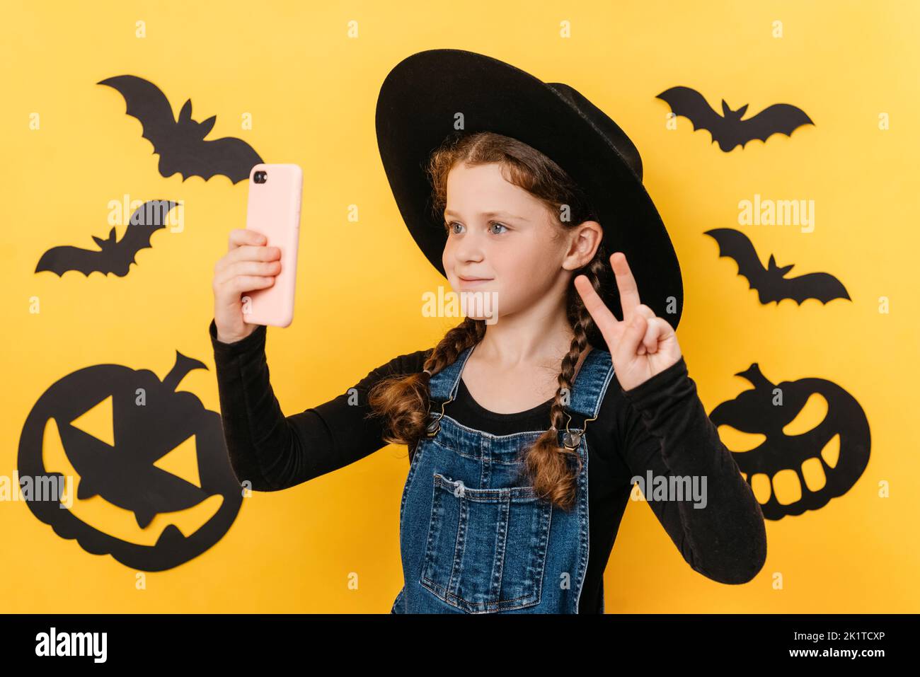 Portrait of pretty little girl kid in hat holding telephone shooting making funky selfies showing v-sign symbol, isolated on yellow color background i Stock Photo