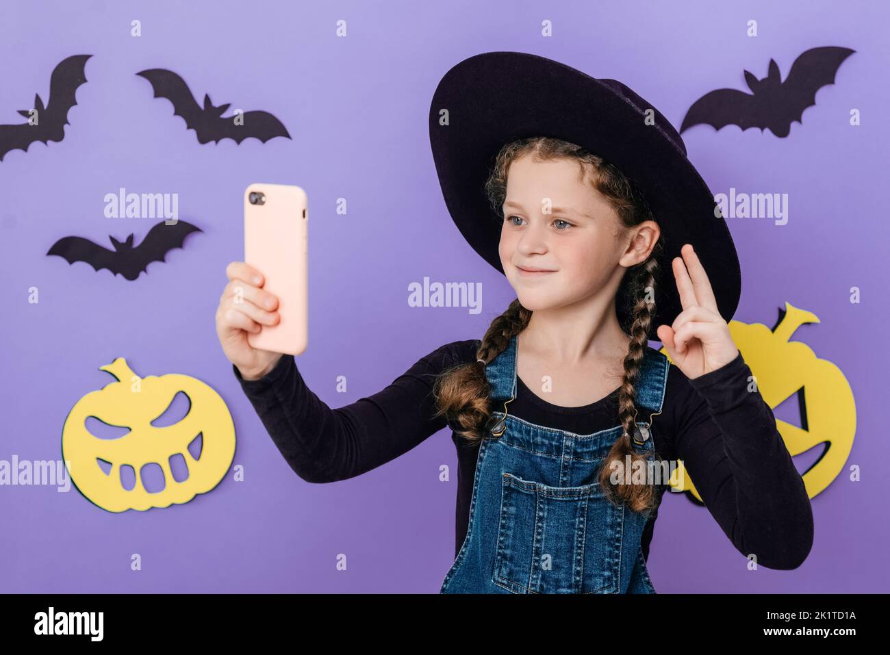 Smiling little girl kid in big hat holding telephone shooting making funky selfies showing v-sign symbol, isolated on purple color background in studi Stock Photo